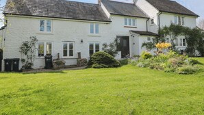 A two-story white house with a sloped roof, a front door, and a small garden with various plants and flowers. - Brook Cottage (Sturminster Newton)