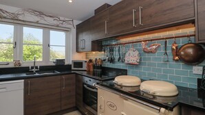 A kitchen with dark wooden cabinets, a white dishwasher, a stove, and a microwave. There is a window with a view of trees outside. - Brook Cottage (Sturminster Newton)