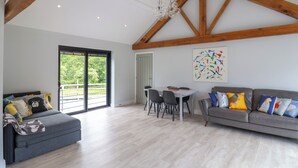 Un'ampia zona giorno con soffitto alto, travi in legno, un divano grigio, un tavolo da pranzo con sedie e una grande finestra che offre vista sul verde.