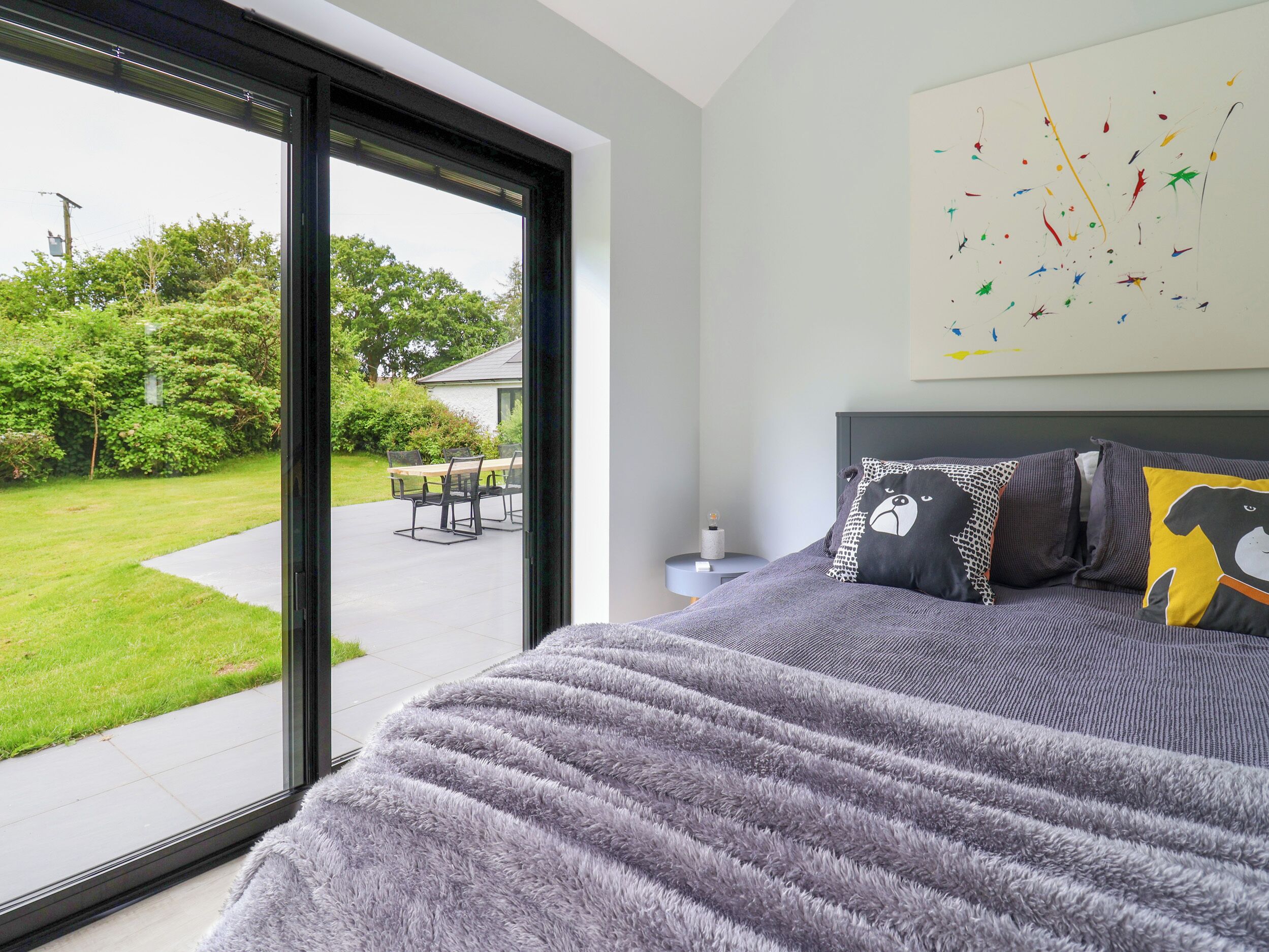 A bedroom with a large window, a bed with grey bedding, and a wall with abstract art.