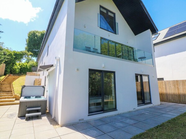 Una casa moderna de dos pisos con balcón, ventanas amplias y un patio con mobiliario exterior.