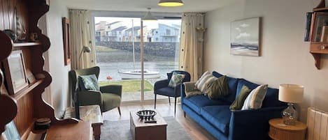 A living room with a blue sofa, a wooden coffee table, a green armchair, and a view of a body of water through large glass doors.