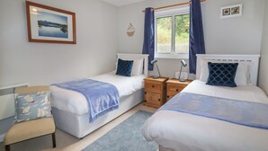 Un dormitorio con dos camas individuales, una mesita de noche con lámpara, una silla, una ventana con cortinas y un cuadro en la pared.