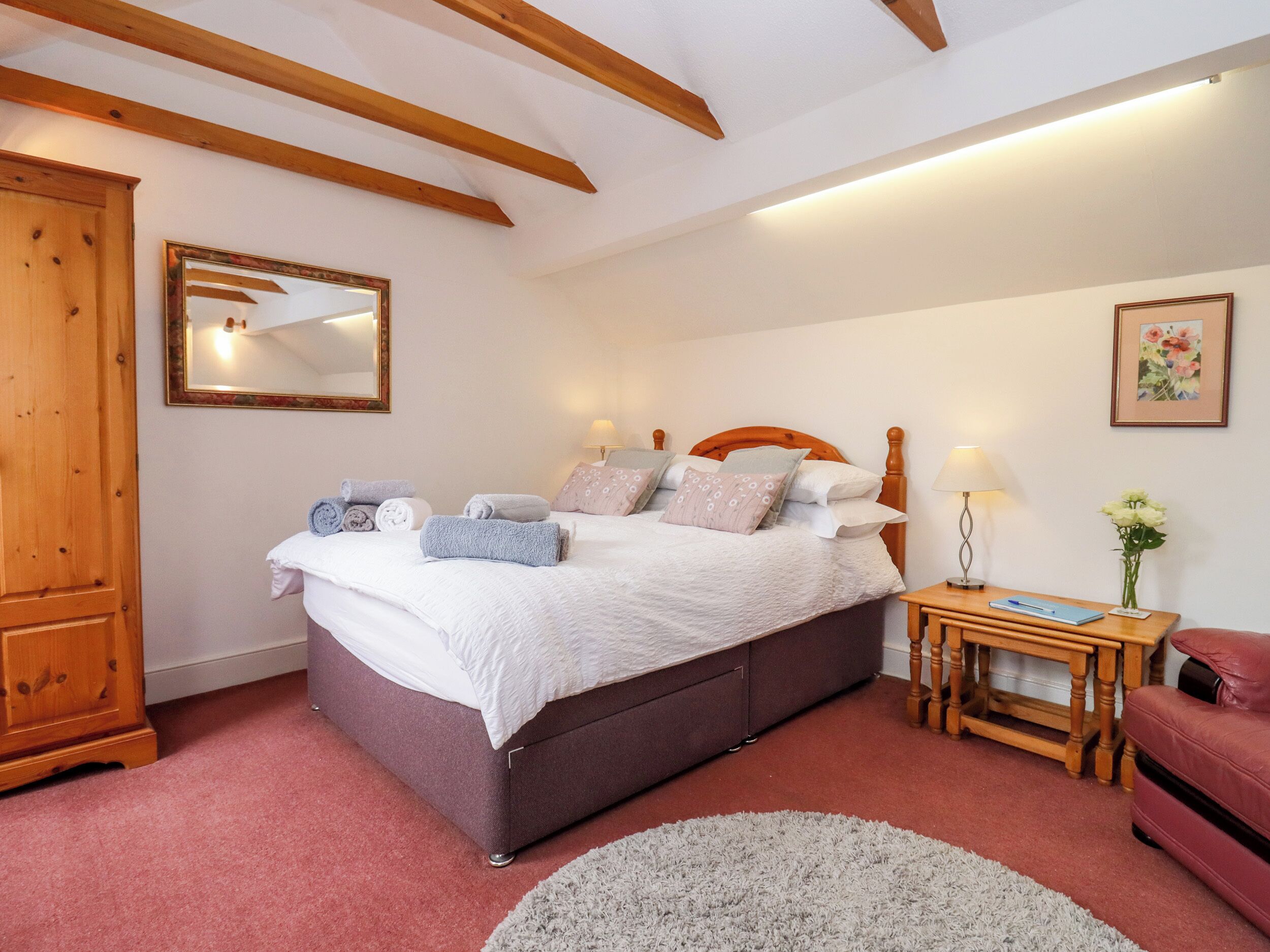 A bedroom with a large bed, wooden beams, a bedside table with a lamp, and a mirror on the wall.