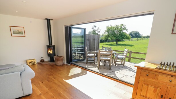 A living room with a sofa, a fireplace, a dining table with chairs, and a view of a green landscape through a glass door. - Little Owl Barn (Worcester)