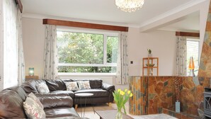 A living room with a leather sofa, a small table, a vase with flowers, a fireplace, and a large window with curtains. - Sea Drift (St Austell)