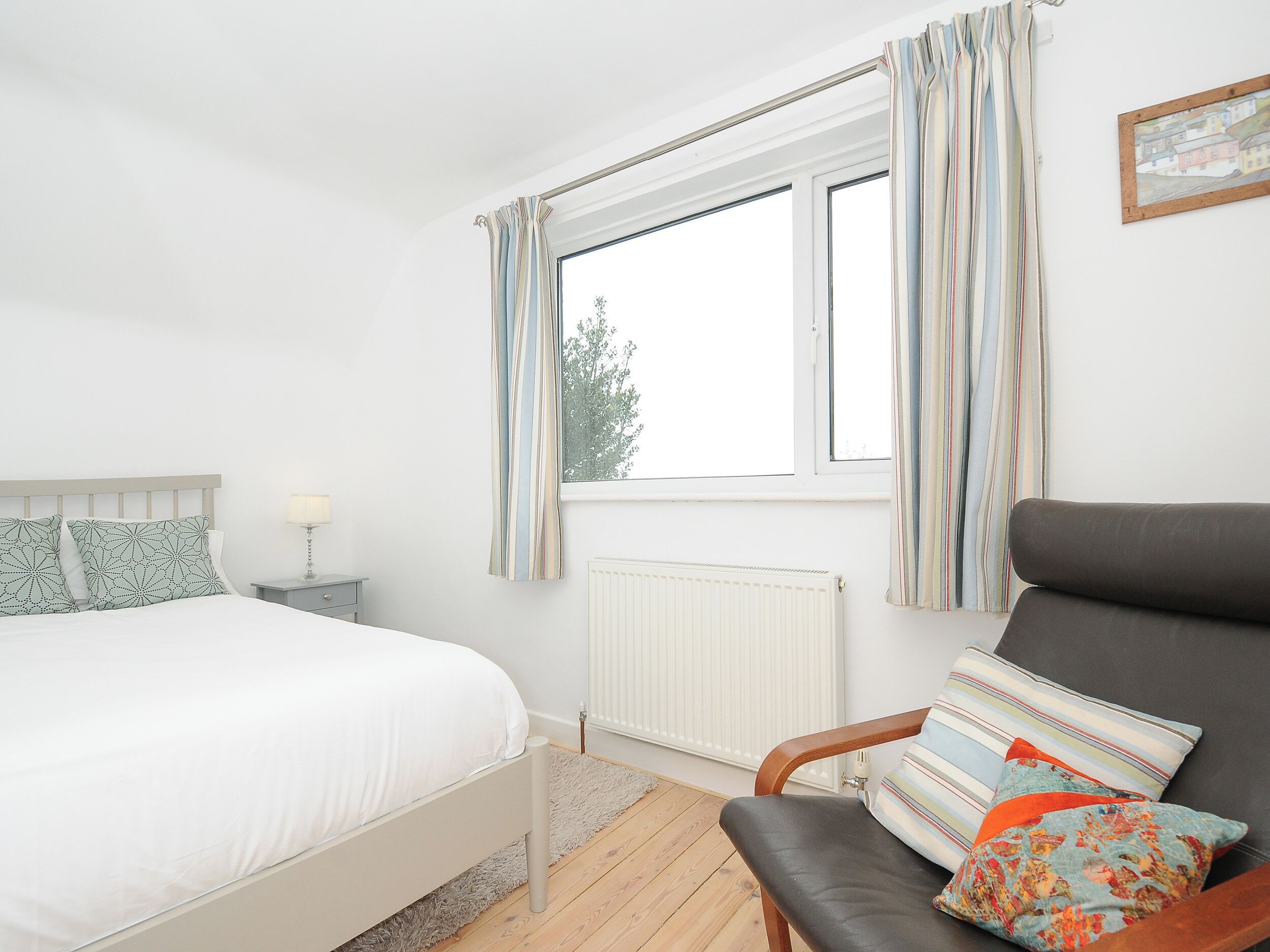 A bedroom with a bed, a chair, a nightstand, a window with curtains, and a radiator.