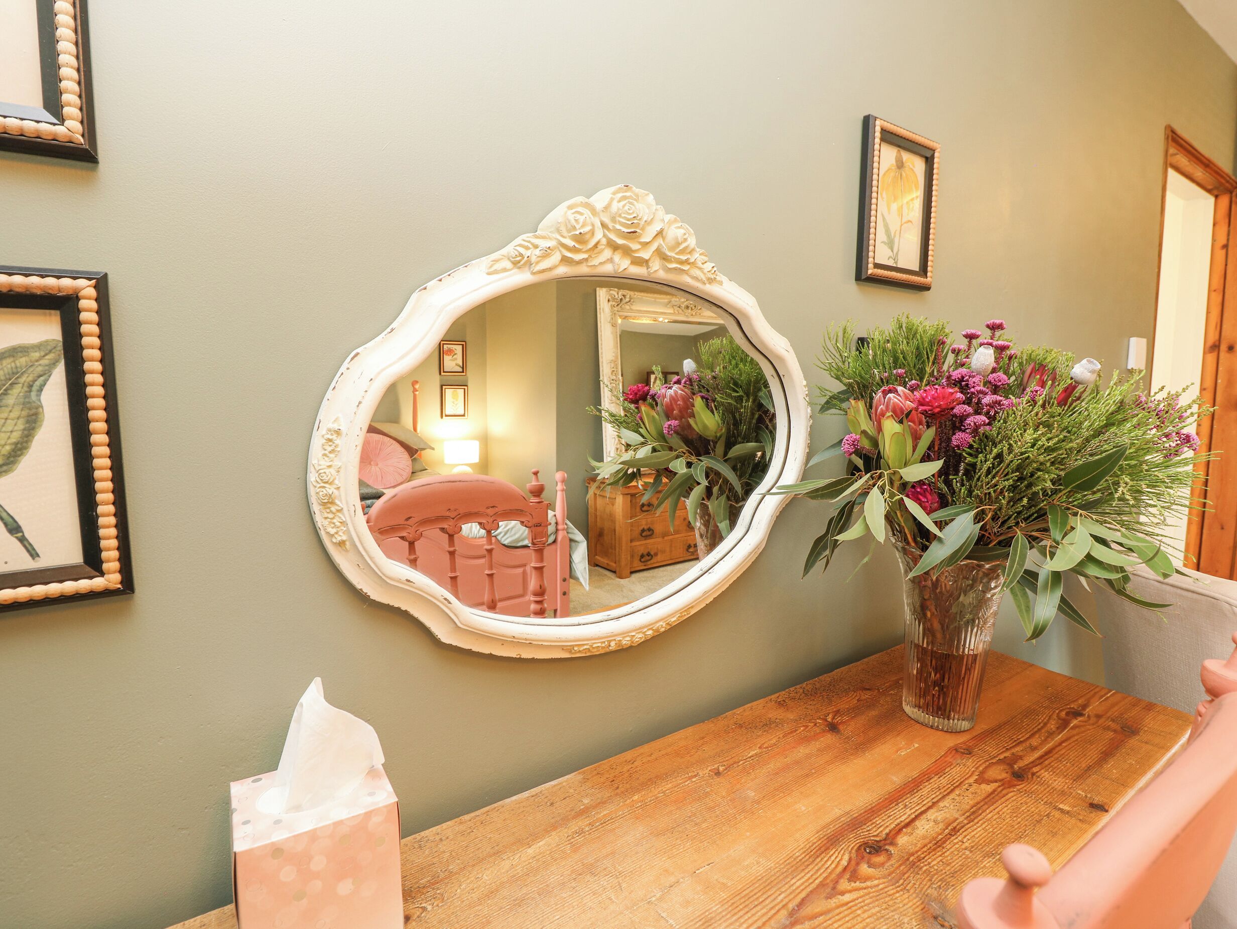 A wooden table with a mirror, a vase with flowers, and a tissue box.