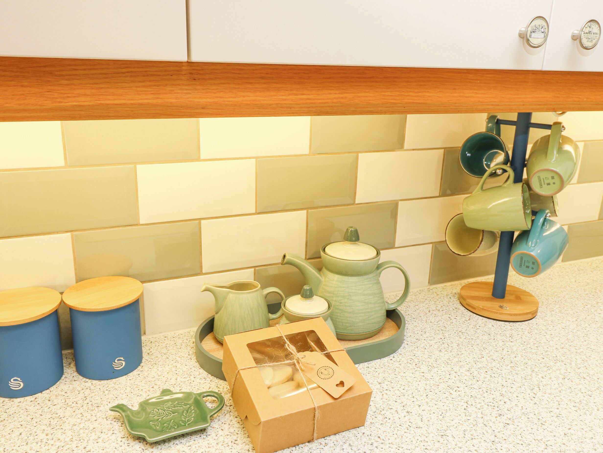 Una cocina con encimera de madera, salpicadero de azulejos y diversos utensilios de cocina, incluyendo un hervidor, tazas y una caja.