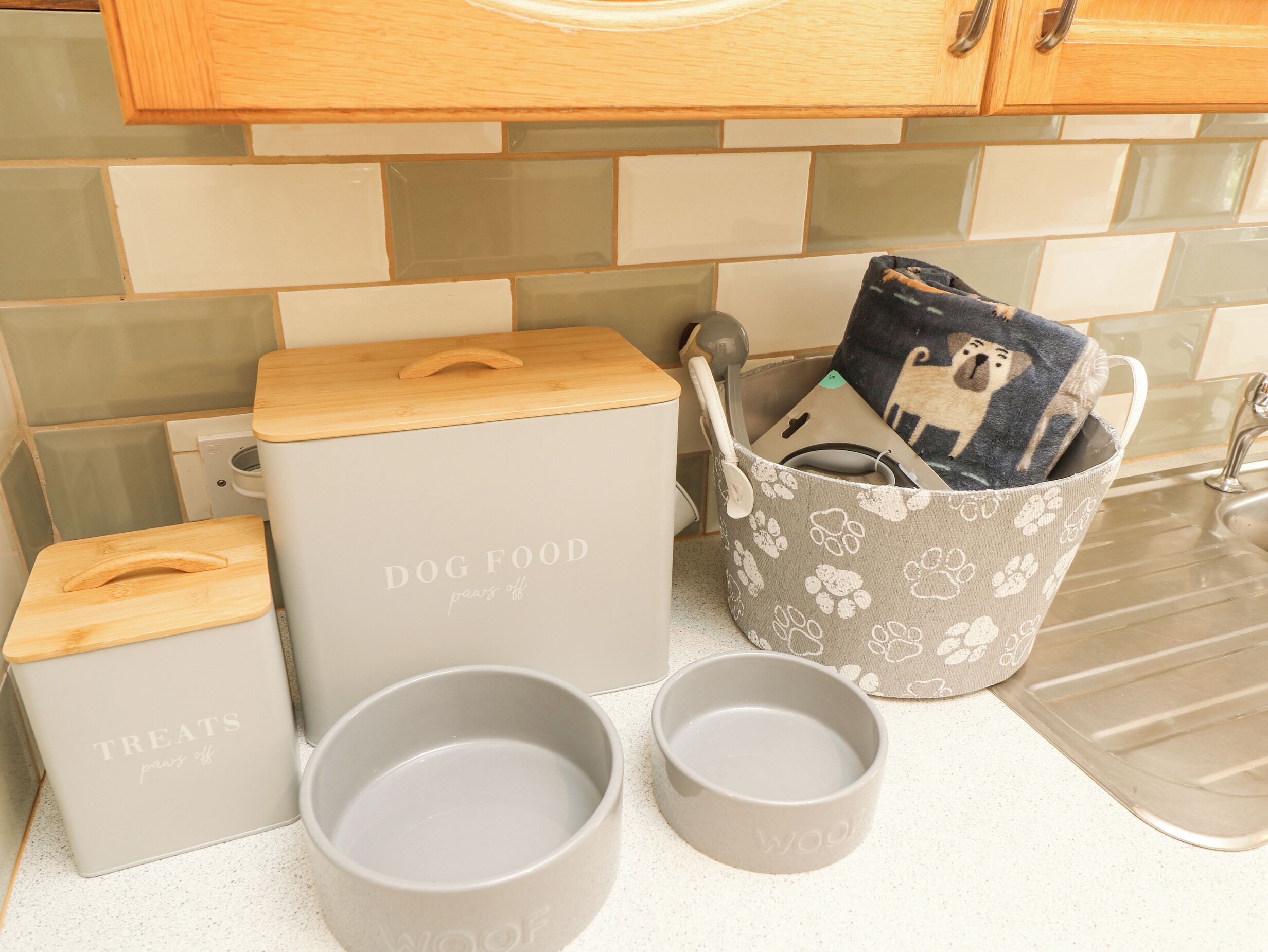 A kitchen counter with storage containers labeled 'DOG FOOD' and 'TREATS,' a grey bucket with a paw print pattern, and two grey mugs with the word 'WOOF' on them.