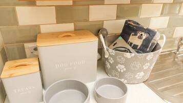 A kitchen counter with storage containers labeled 'DOG FOOD' and 'TREATS,' a grey bucket with a paw print pattern, and two grey mugs with the word 'WOOF' on them.