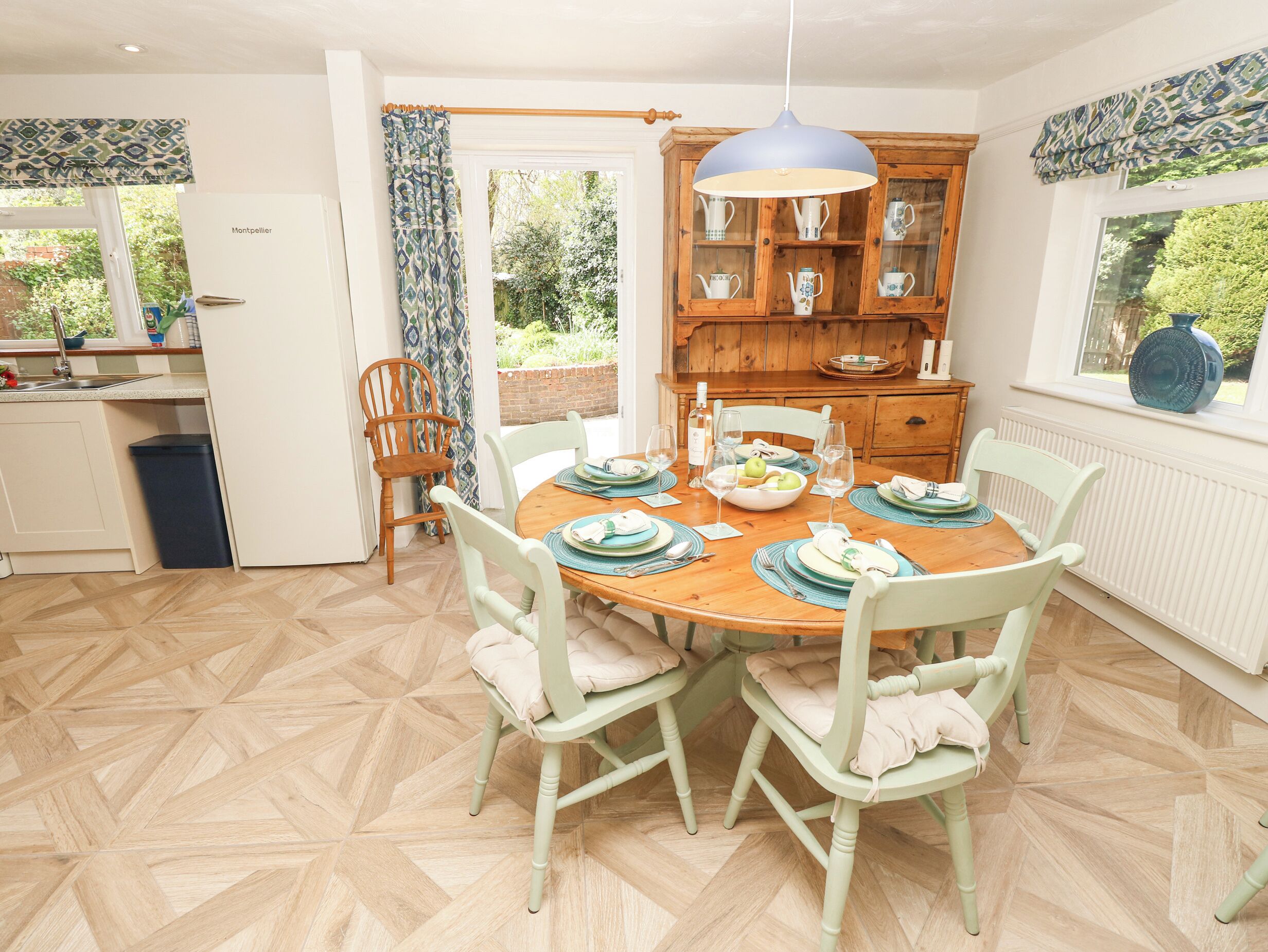 A dining area with a wooden table set for four, surrounded by light green chairs with cushions. A wooden cabinet with glass doors is visible in the background.