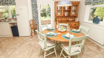 A dining area with a wooden table set for four, surrounded by light green chairs with cushions. A wooden cabinet with glass doors is visible in the background.