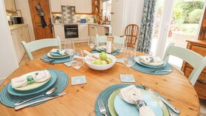 A dining area with a wooden table set for four, surrounded by light-colored chairs. A bowl of green apples and wine glasses are on the table. - Horseshoe Cottage (Romsey)