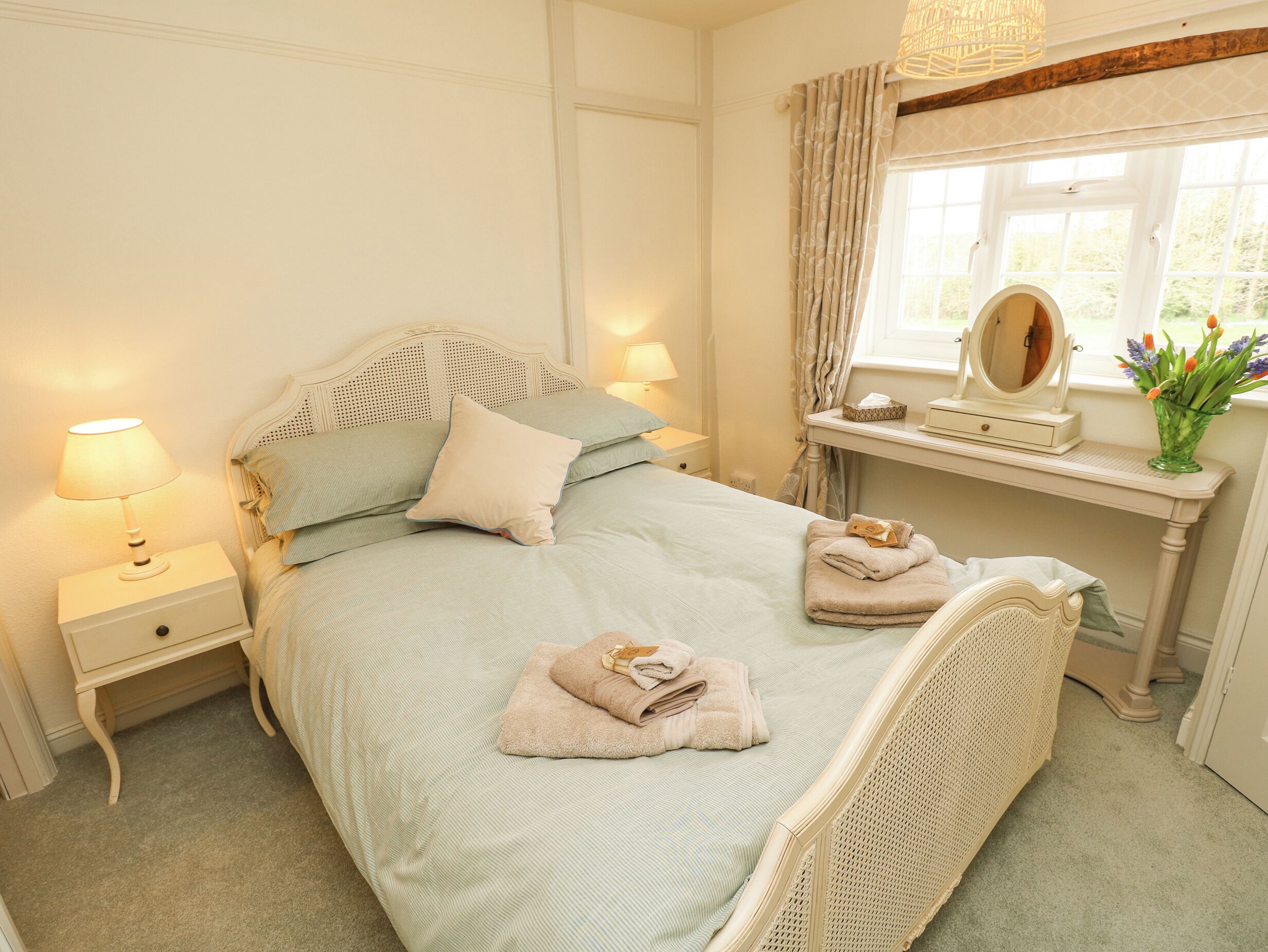A neatly made bed with pillows and folded towels, a bedside table with a lamp, a vanity with a mirror, and a window with curtains.