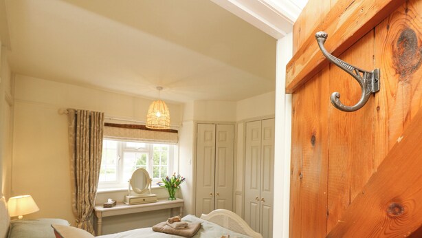 A bedroom with a bed, a bedside table with a lamp, a window with curtains, a mirror, and a wooden door with a metal handle. - Horseshoe Cottage (Romsey)