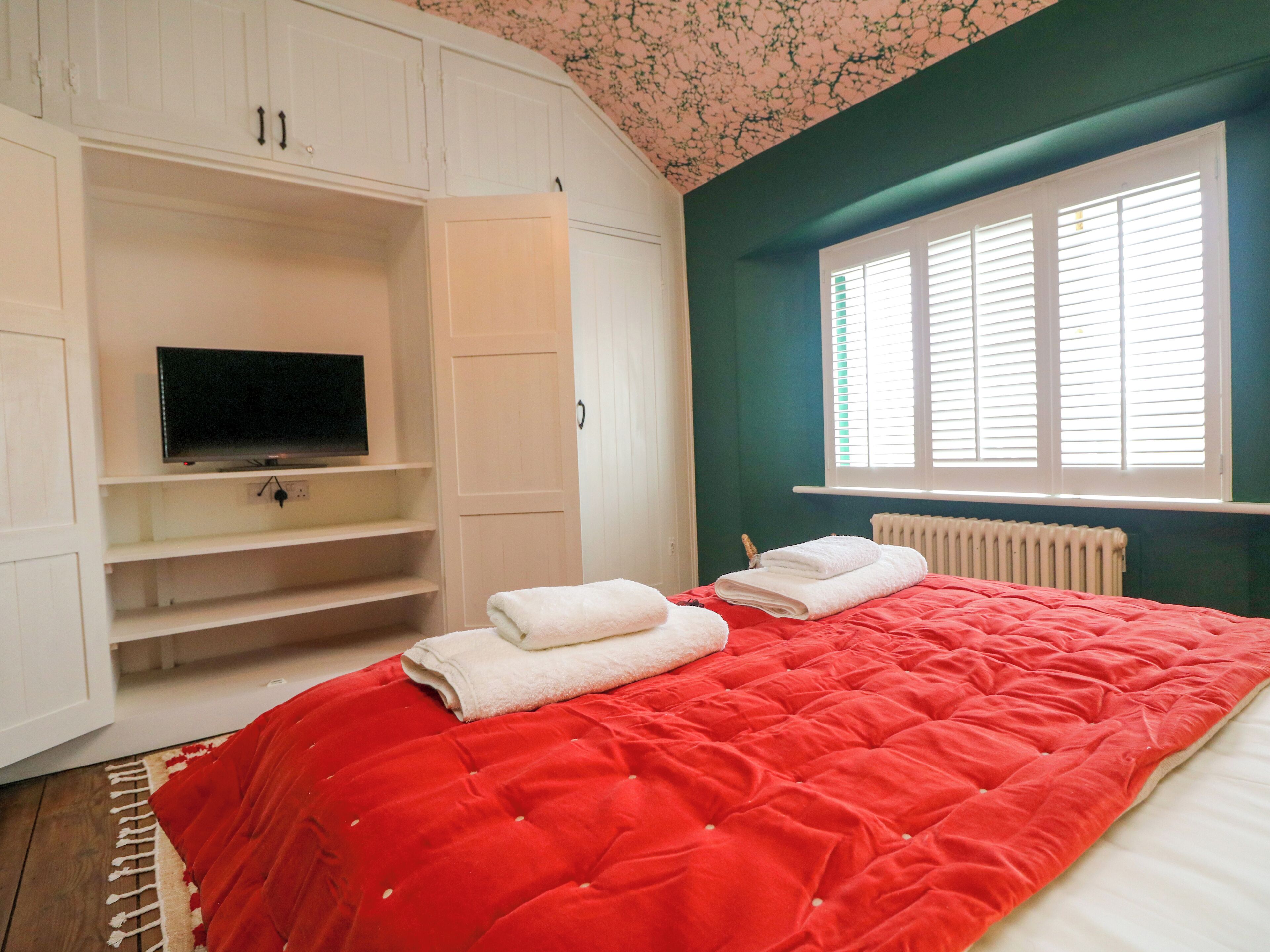 A bedroom with a red bedspread, a television on a white shelf, and a window with blinds.