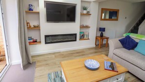 Una sala de estar con televisor de pantalla plana, chimenea, estantes con objetos decorativos, una mesa de centro con un cuenco y un libro, y un sofá con cojines.