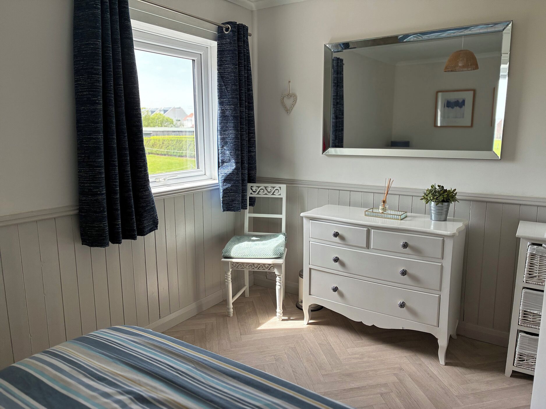 Un dormitorio con cama, cómoda, silla, ventana con cortinas, espejo y vistas al exterior.