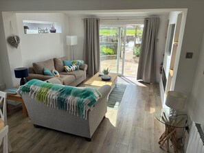 A living room with a sofa, a coffee table, a lamp, and a view of the outdoors. - Lavender Cottage (Chathill)