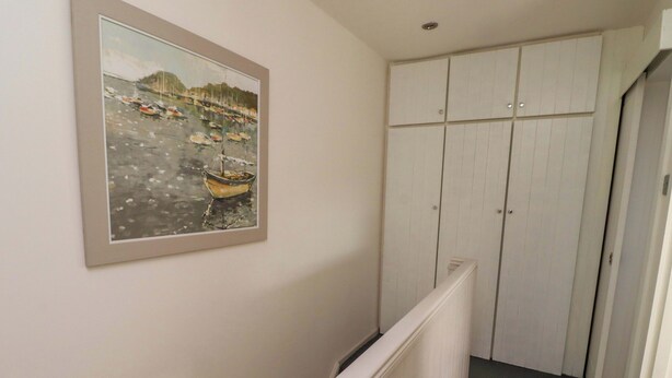 A hallway with a framed painting of boats on the wall, a white radiator, and a set of white cabinets. - Lavender Cottage (Chathill)