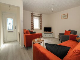 Un salón con sofás naranja, un televisor sobre un mueble de madera, una ventana con cortinas estampadas y una puerta con panel de vidrio.