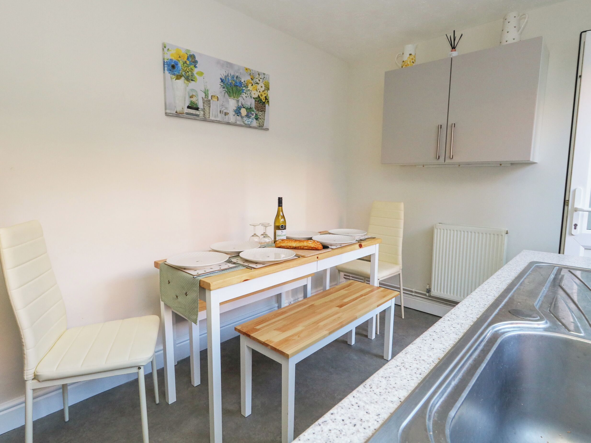 A small, modern kitchen with white cabinetry, a wooden table, and a bench. There is a painting of flowers on the wall, and a radiator is visible.