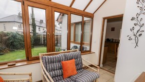Una habitación acogedora con un sofá y sillón de mimbre, amplias ventanas con vistas a un jardín y casas, y una pared blanca con un sencillo adorno de hoja.