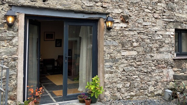 A stone building with a glass door entrance, potted plants, and a metal watering can on the ground. - Eden Vale Cart House (Broughton in Furness)
