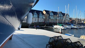 Un port de plaisance avec des bateaux amarrés, un bâtiment doté de nombreuses fenêtres et la coque d’un grand bateau au premier plan.