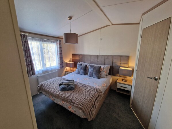 A bedroom with a bed, bedside lamps, a radiator, and a window with curtains. - Ladybird Lodge (Carnforth)