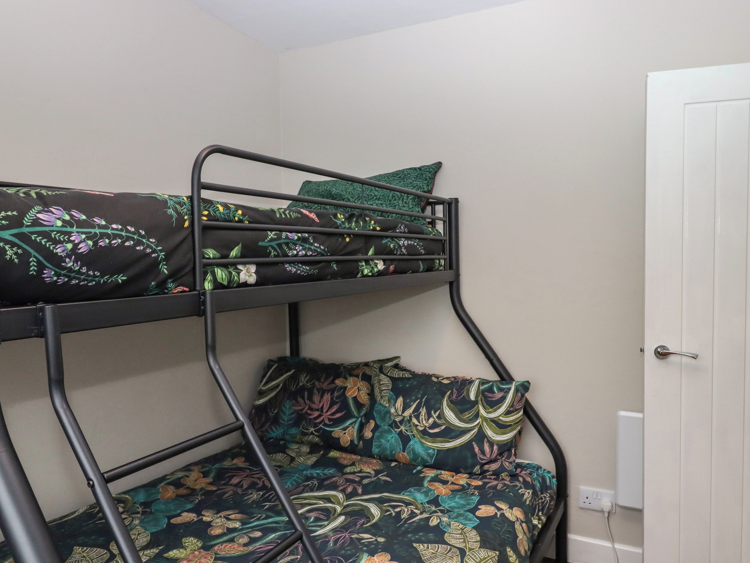 Una cama litera con ropa de cama estampada en motivos florales en una habitación con una puerta blanca y un enchufe en la pared.