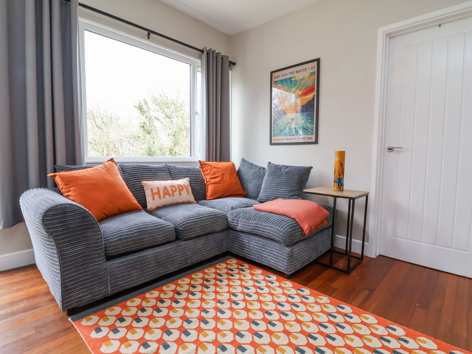 A modern living room with a grey sectional sofa, orange cushions, a patterned rug, a small side table, and a framed poster on the wall.