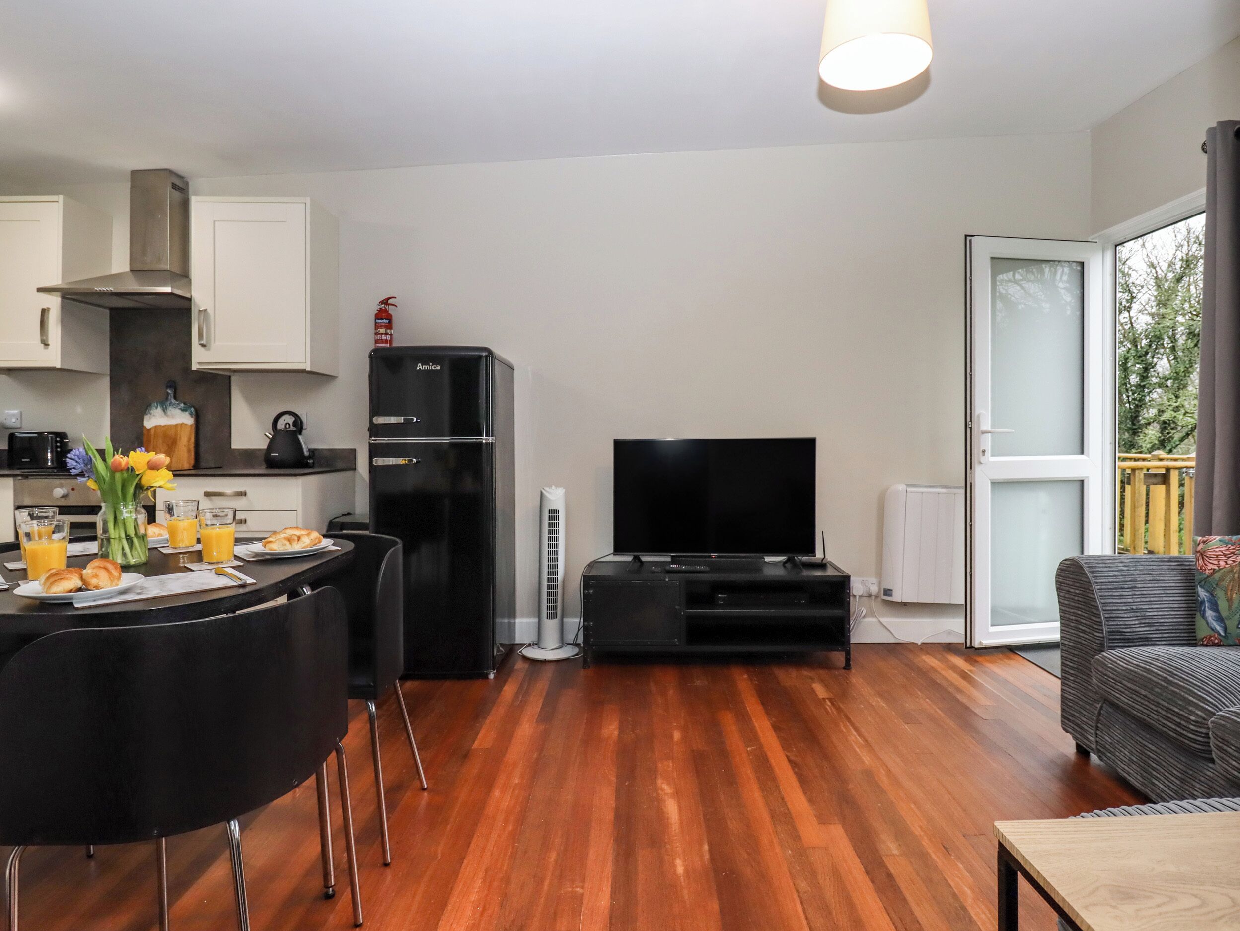 A modern living room with a flat-screen TV, a black refrigerator, and a dining area with a table set for two.