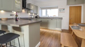 A modern kitchen with grey cabinets, a central island with bar stools, a window, and a wooden dining table. - 2 South Street (Wigton)
