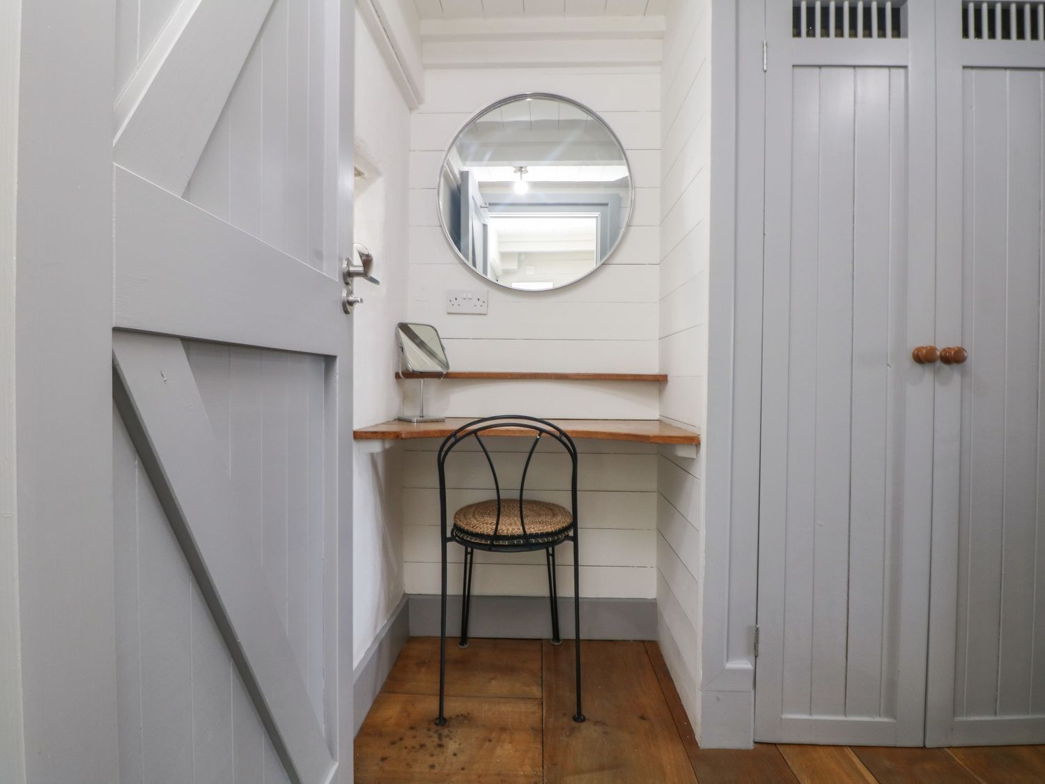 Un couloir étroit doté d’un plancher en bois, d’une chaise en métal noir, d’une étagère en bois, d’un miroir rond et d’une porte blanche avec une poignée.