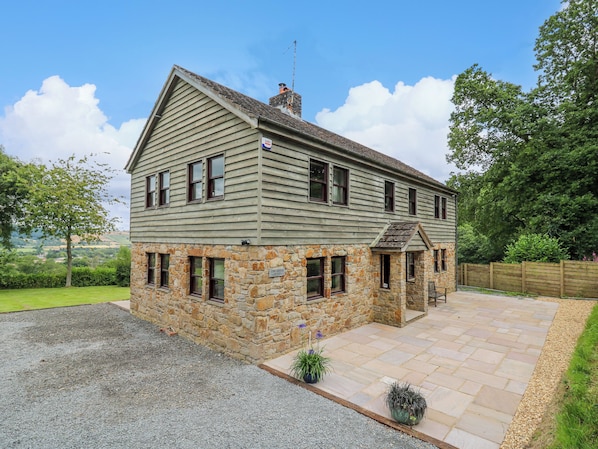 A two-story stone cottage with a stone foundation and wooden siding, a paved driveway, and a view of the surrounding landscape. - Oak Apple Cottage (Presteigne)