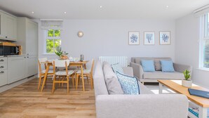 Una sala moderna con un sofá gris, una mesa de comedor de madera y una zona de cocina con armarios blancos y un microondas.