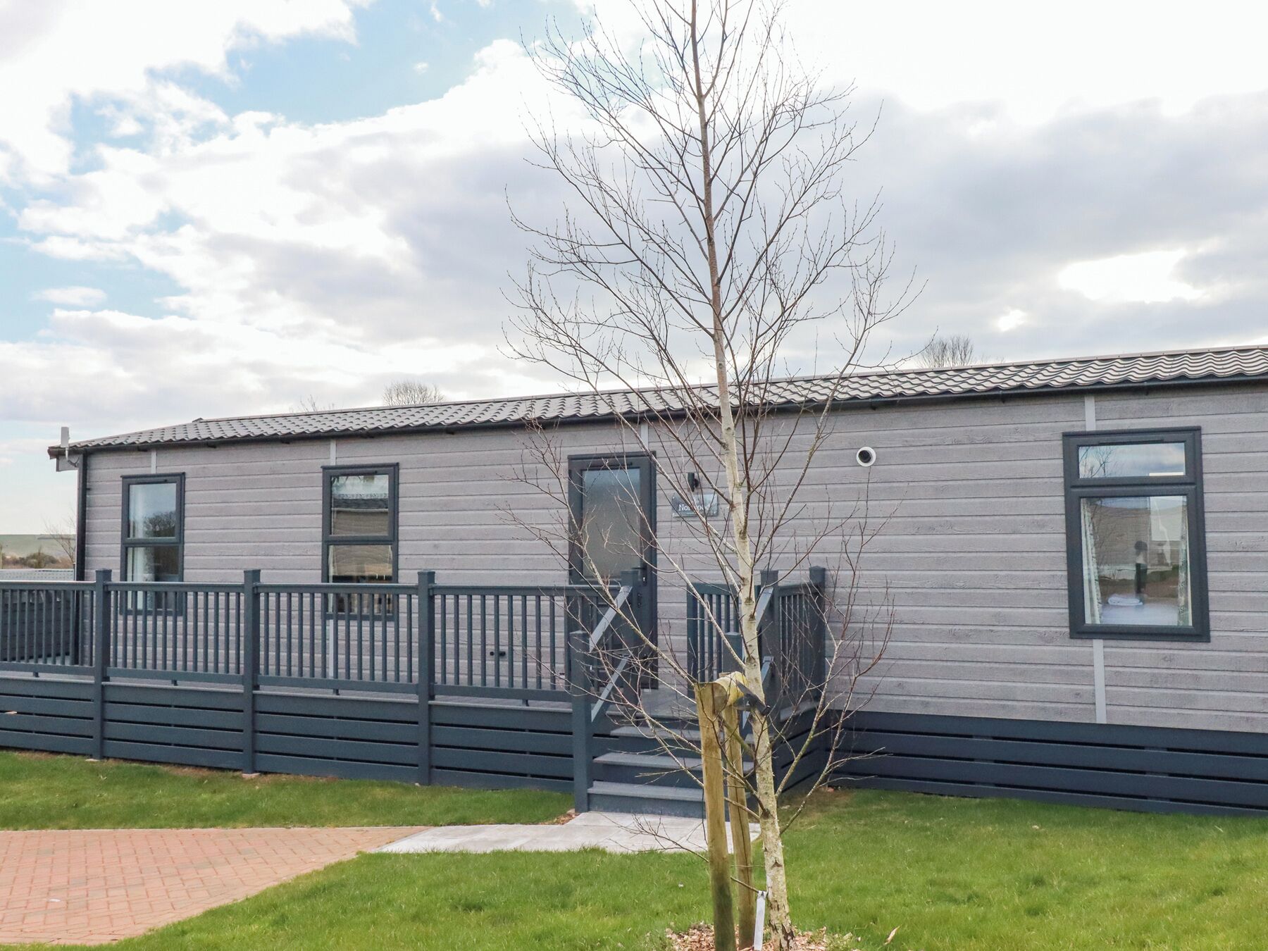 A modern, single-story mobile home with a grey exterior and black trim. It features a front door, two windows, and a small porch area.