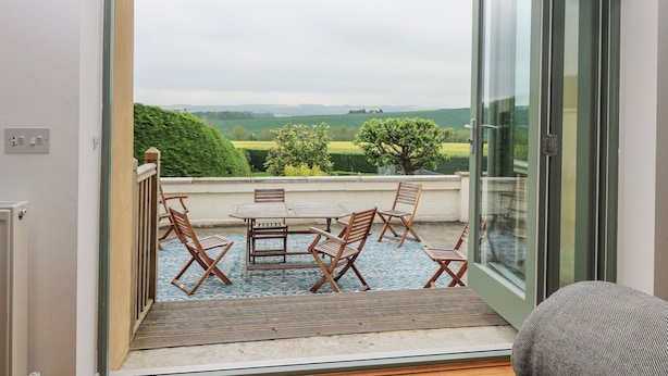 Un balcon avec une table et des chaises, une vue sur des champs verdoyants et un ciel nuageux.