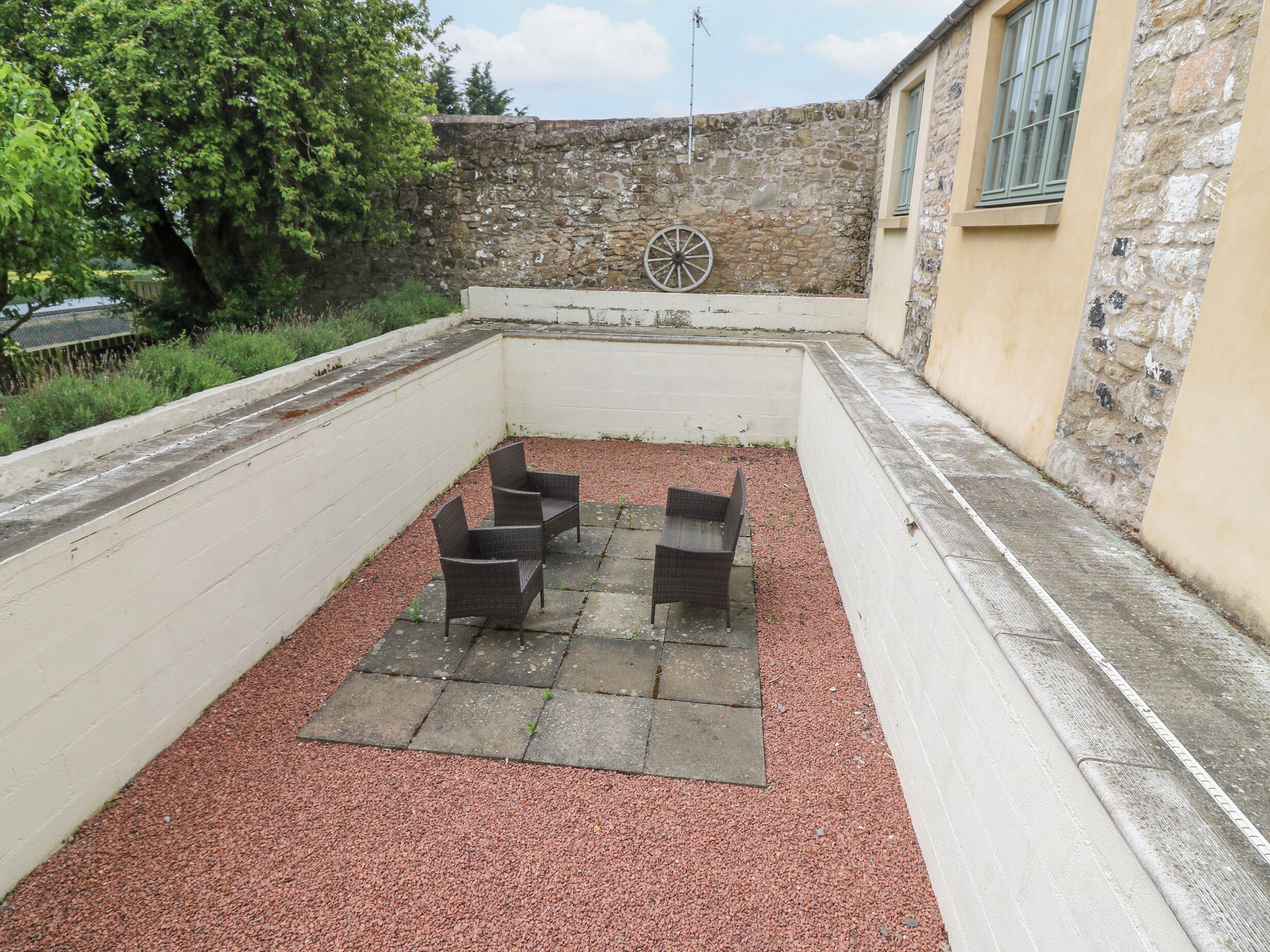 An outdoor seating area with four wicker chairs and a table, surrounded by a stone wall and a building with a window.