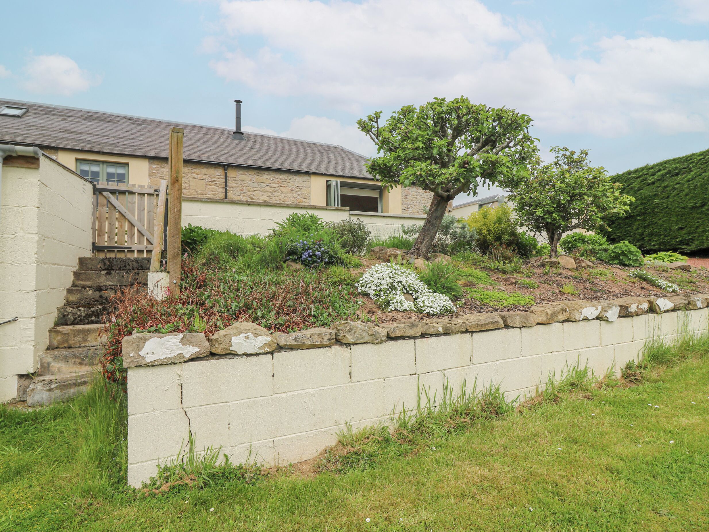 A house with a stone facade, a wooden staircase, and a garden with various plants and a tree.