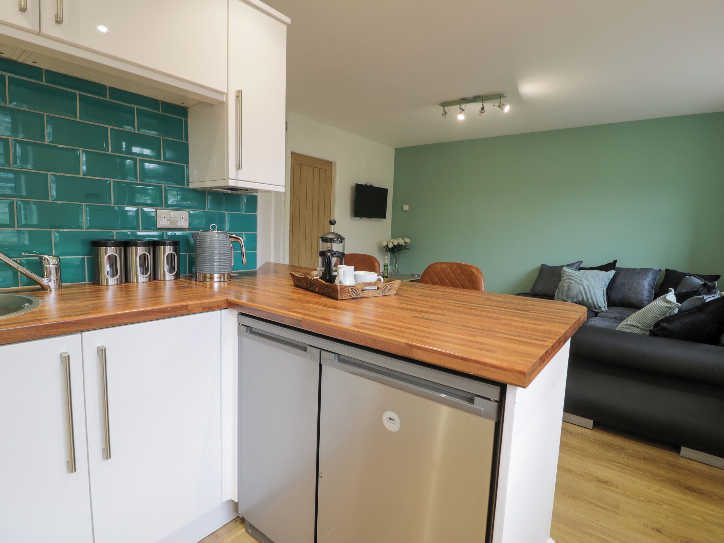 A modern kitchen with white cabinets, a wooden countertop, and a dishwasher. There is a teal backsplash and a dining area with a dark sofa and a coffee table.