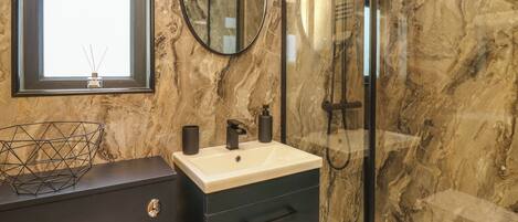 A modern bathroom with a marble wall, a round mirror, a black vanity, and a glass-enclosed shower.