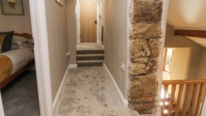A narrow hallway with carpeted stairs leading to a wooden door. A stone pillar is on the right, and a bed with pillows is visible on the left. - High Winder House (Penrith)