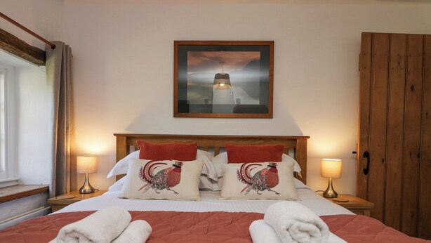 A hotel room with a large bed, two red pillows with a rooster design, a framed artwork above the bed, and two bedside lamps. - High Winder House (Penrith)