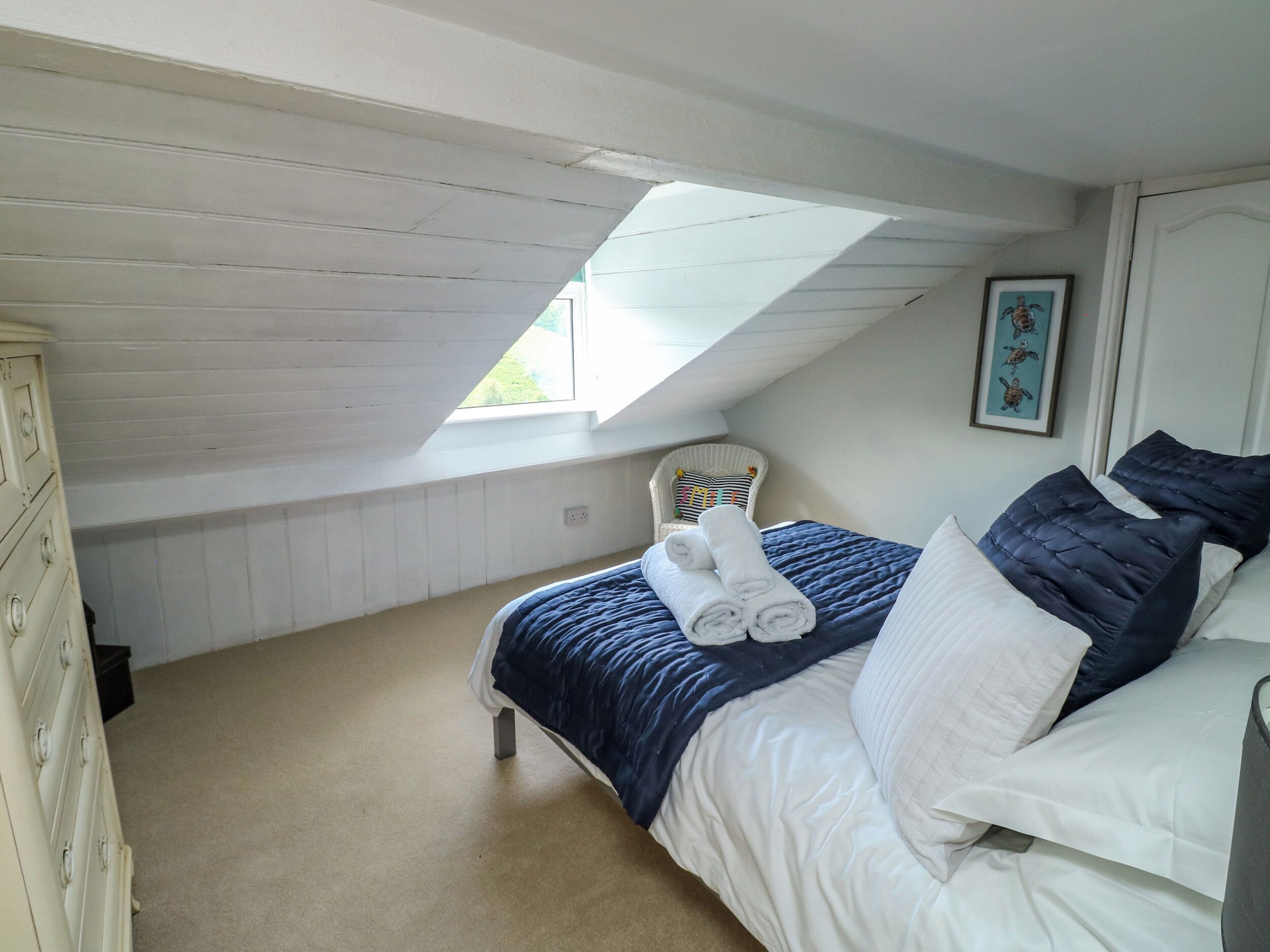 Une chambre avec un plafond en pente, un lit avec des serviettes enroulées, une chaise et un tableau encadré au mur.