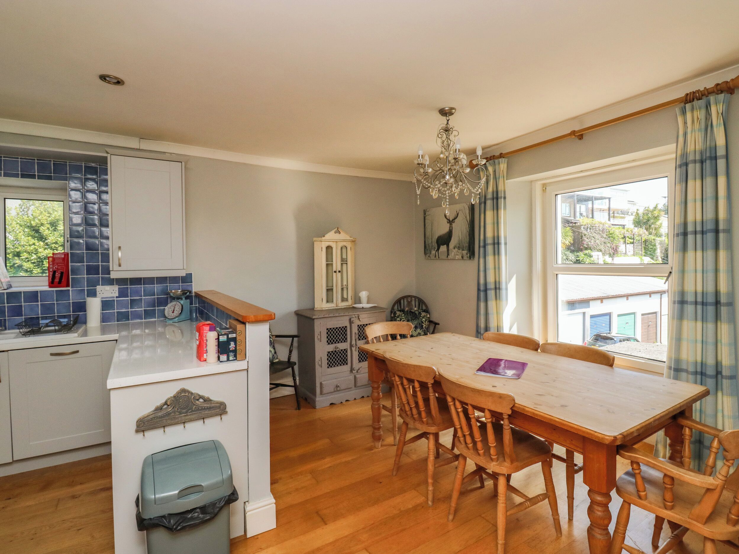 A kitchen with wooden cabinets, a dining table with chairs, a chandelier, and a view of the outside through a window.