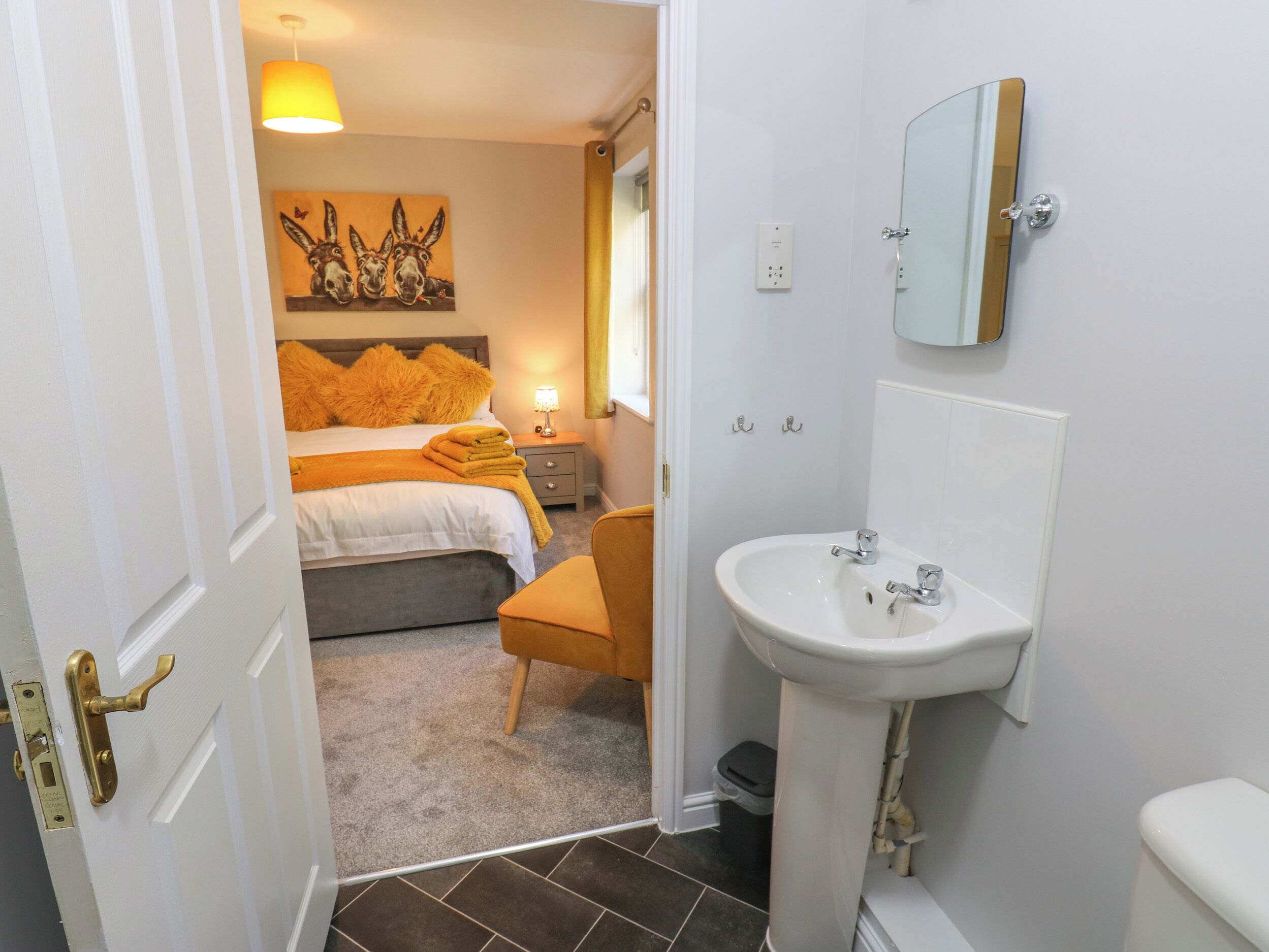 A bedroom with a bed, a bedside table with a lamp, a bathroom with a sink and mirror, and a door leading to another room.