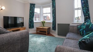A living room with a flat-screen TV, grey sofas, a small wooden table, and a vase of flowers. - The Chase - 30 Ayr Avenue (Catterick Garrison)